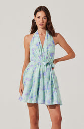 A woman is wearing a sleeveless, halter-neck dress with a waist tie. The dress has a light, flowing fabric with a pastel watercolor pattern in shades of blue, green, and purple.